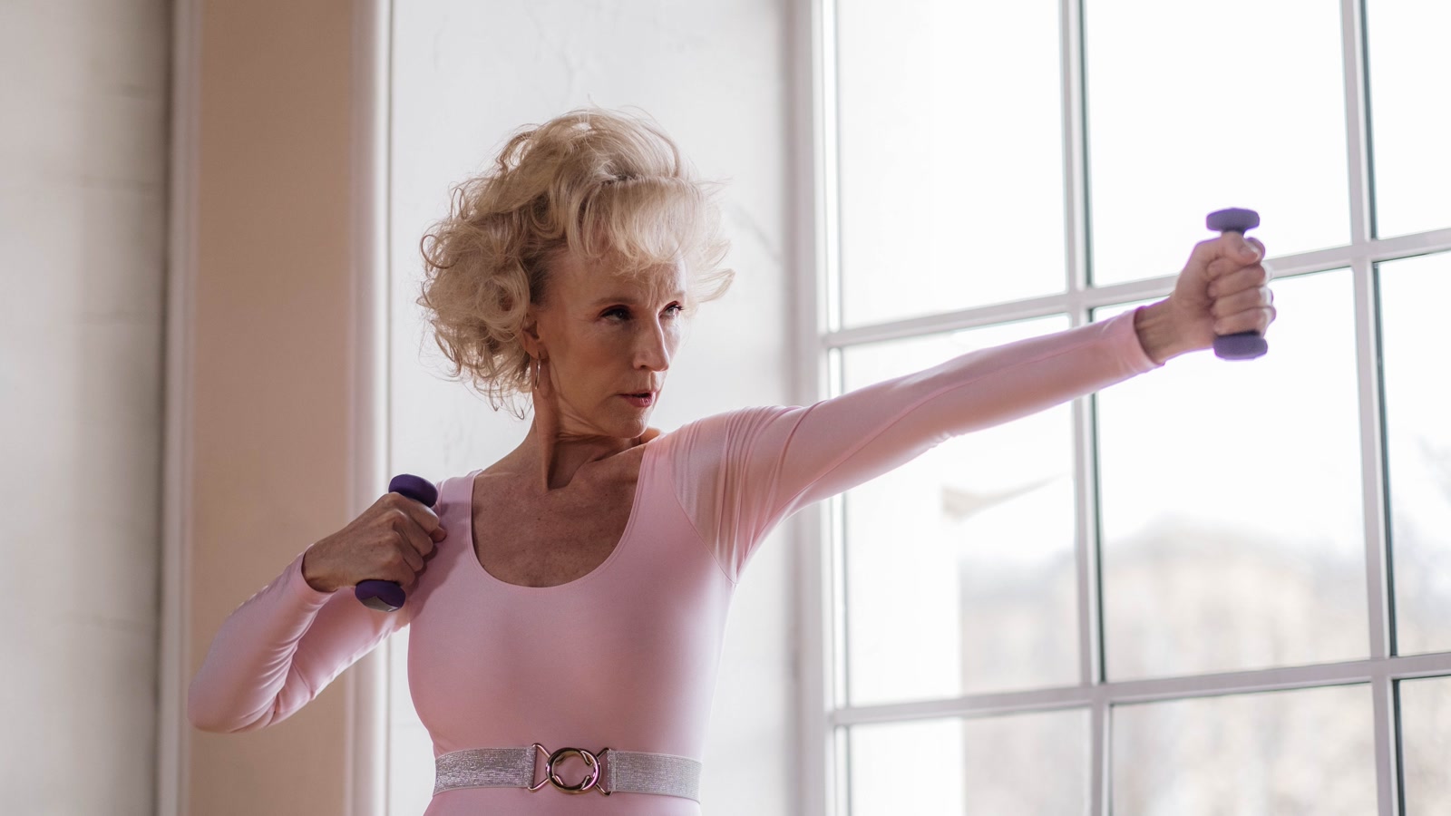 Woman over 40 doing strength exercises with purple dumbbells near a bright window