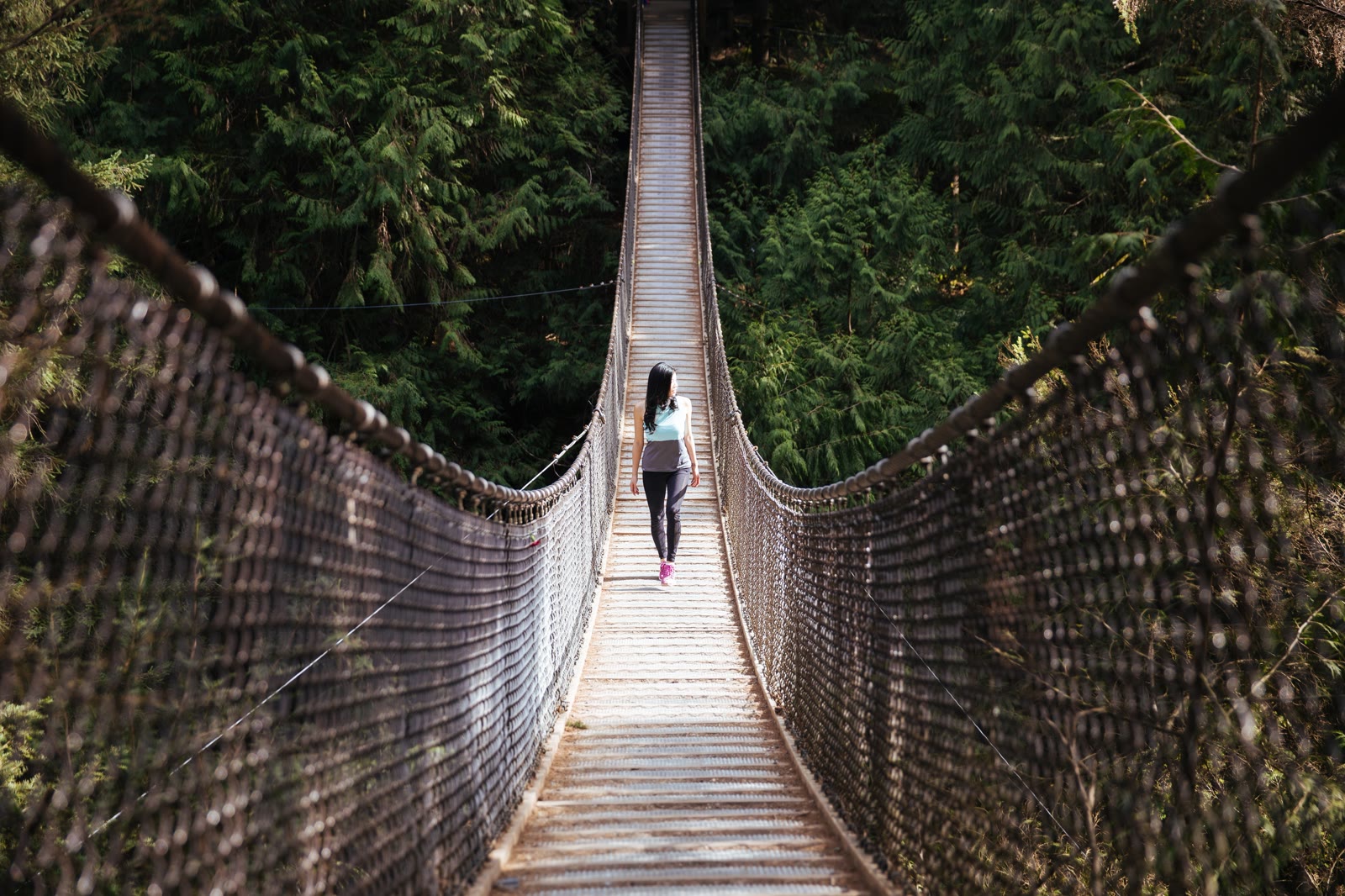 Woman walking away from the camera across a suspension bridge in a green forest